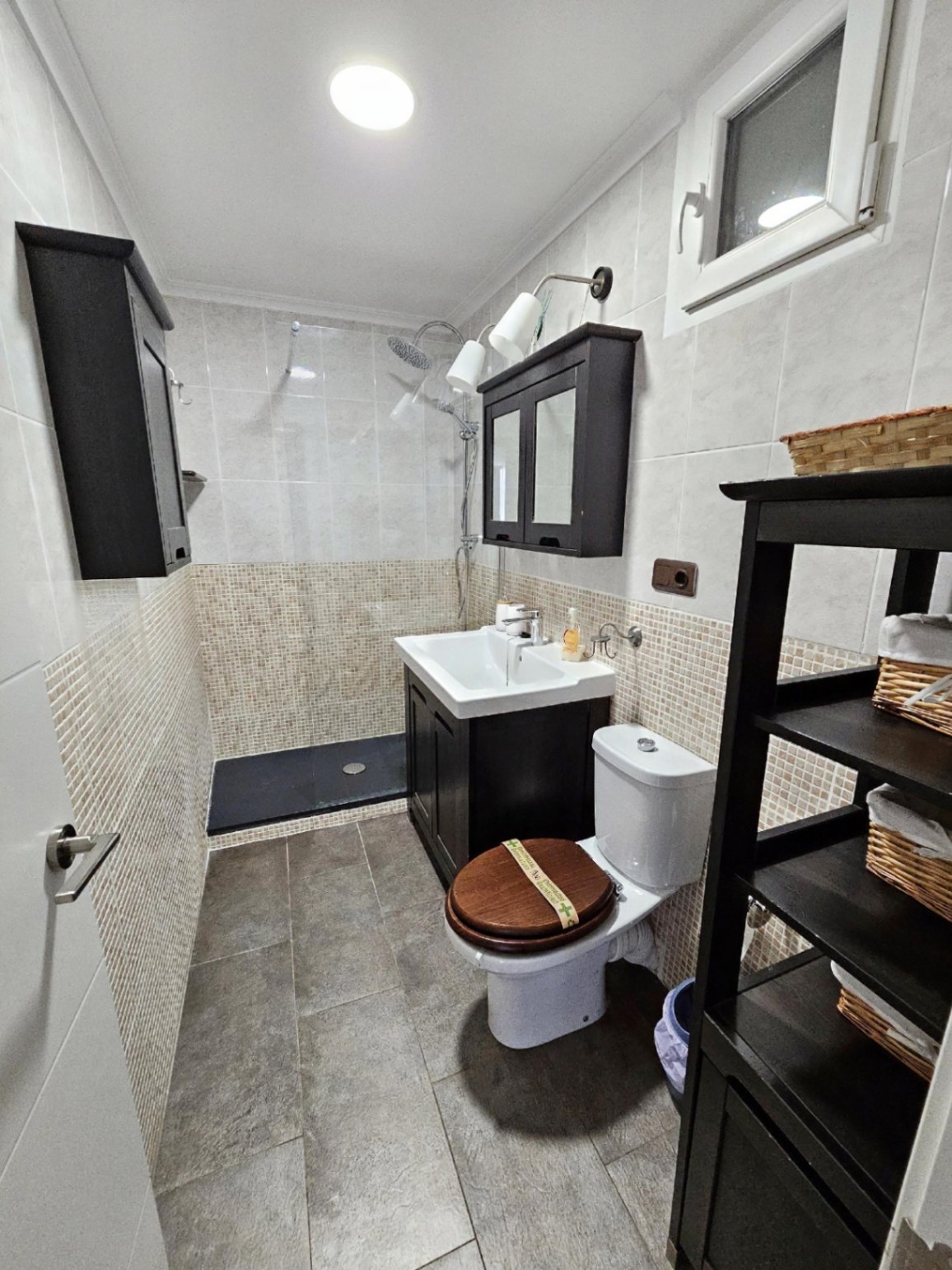 Modernisiertes Badezimmer mit begehbarer Dusche, Mosaikfliesen und dunklen Holzmöbeln, die einen eleganten Kontrast zum weißen Sanitär bilden.