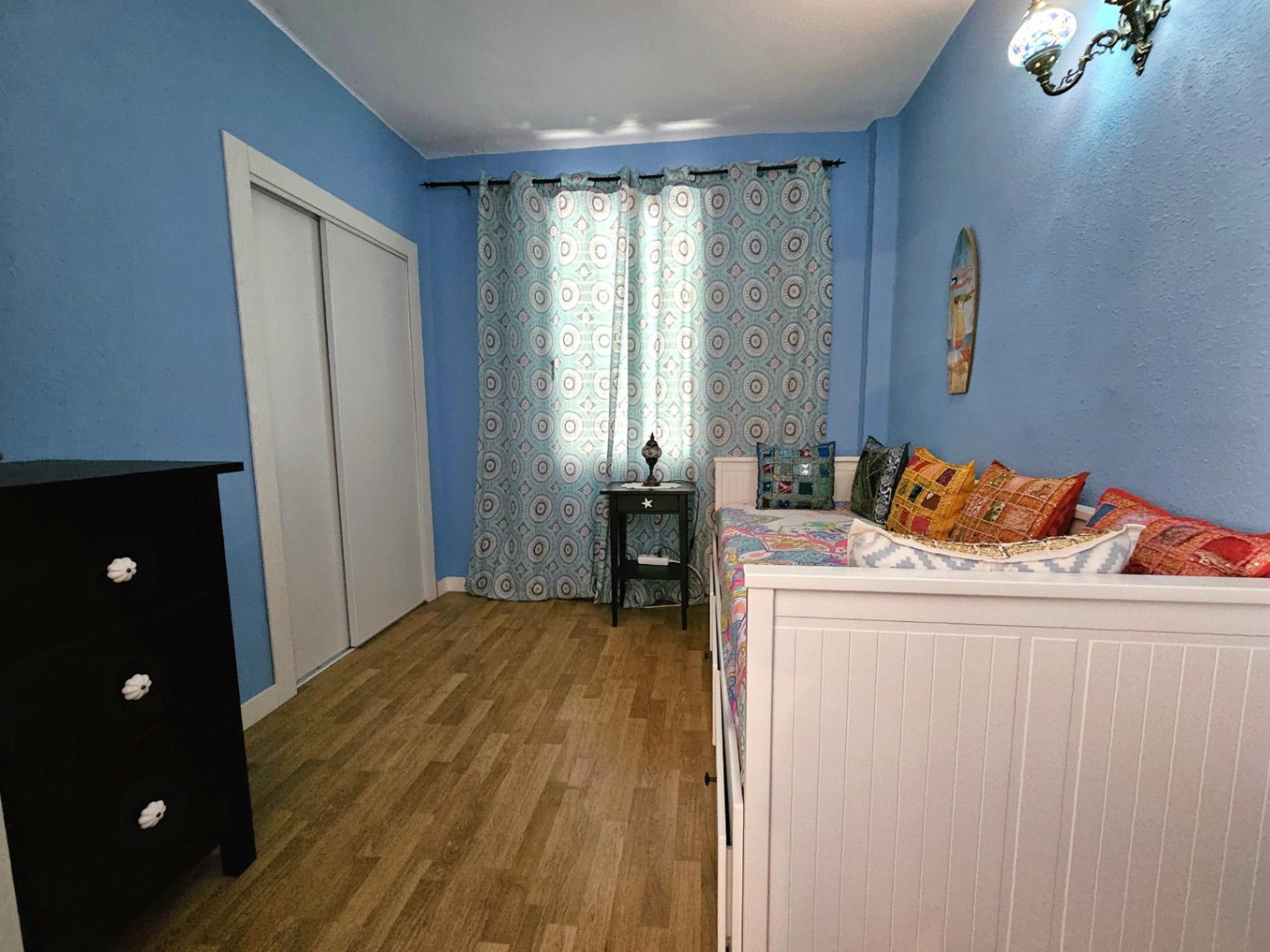 Schlafzimmer mit hellblauen Wänden, Parkettboden und weißem Einbauschrank. Es verfügt über ein weißes Tagesbett mit bunten Kissen und eine dunkle Kommode.