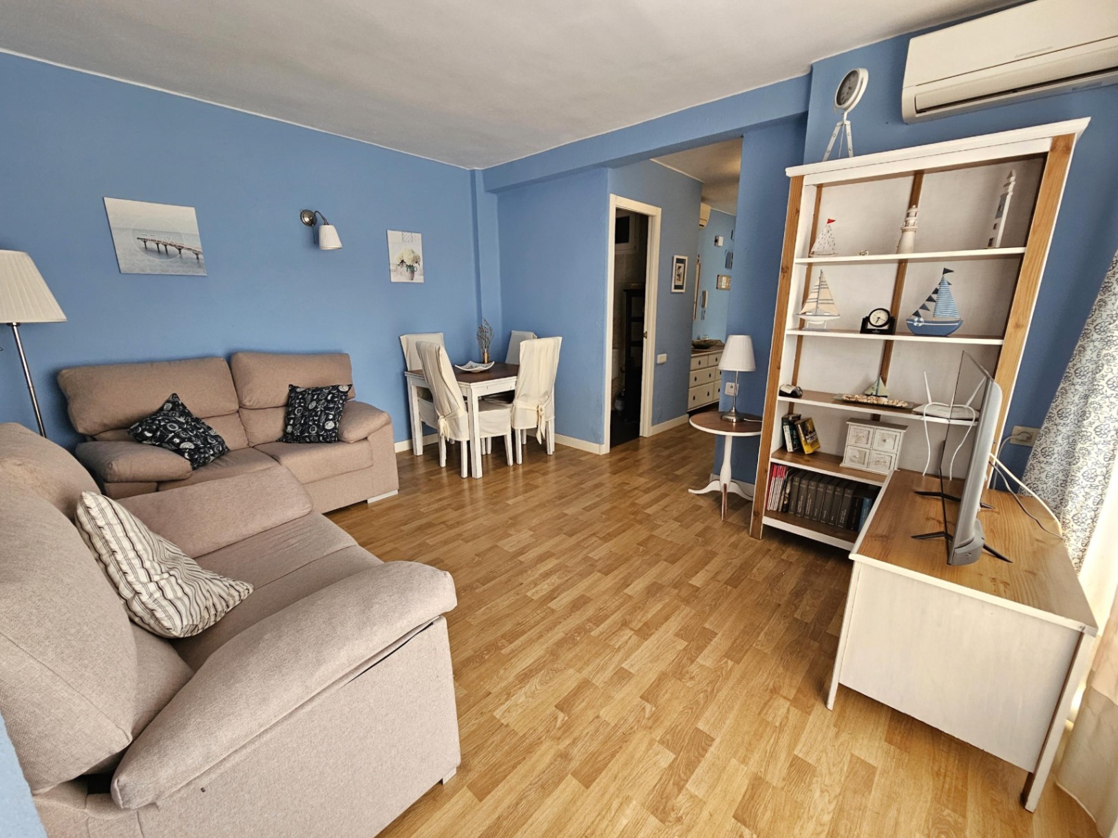 Wohn- und Essbereich mit blauen Wänden, Parkettboden und beigem Sofa. Ausgestattet mit einem weißen Holzregal und einer Essgruppe für vier Personen.