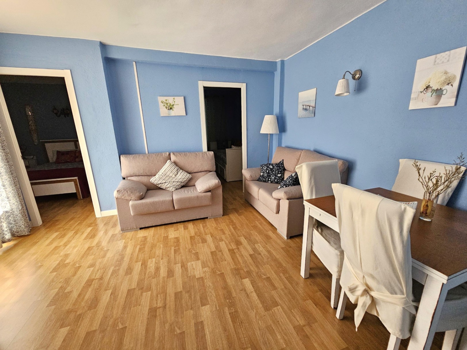 Wohn- und Essbereich mit blauen Wänden, Parkettboden und modernen Möbeln. Der Raum bietet zwei neutrale Sofas und einen Essplatz neben den Zugängen zu den Schlafzimmern.