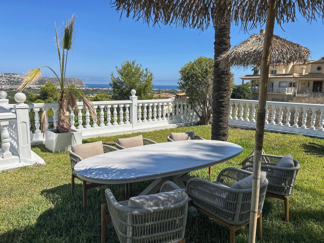 Außen-Essbereich auf einer Rasenfläche mit ovalem Tisch und Flechtstühlen, begrenzt durch eine weiße Balustrade mit weitem Blick auf das Meer und das Tal.