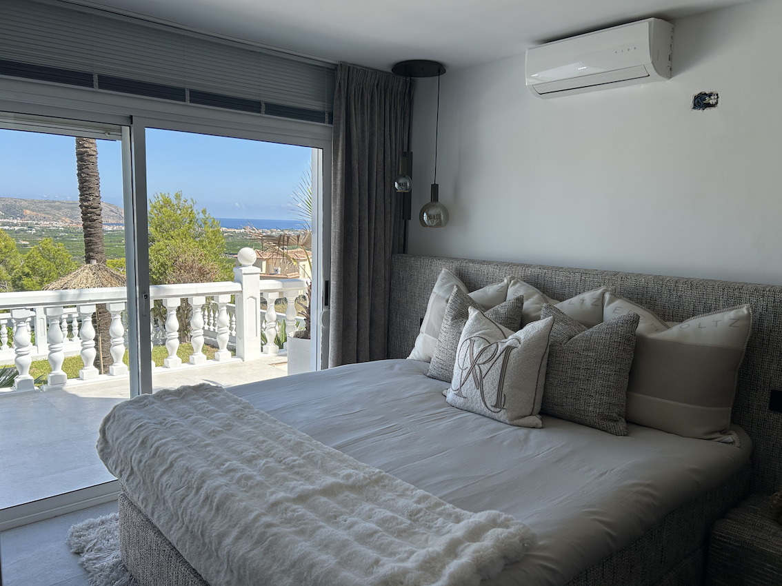 Hauptschlafzimmer mit gepolstertem Kopfteil und Klimaanlage. Die Glasschiebetür führt auf eine private Terrasse mit weitem Blick auf das Meer und das Tal.