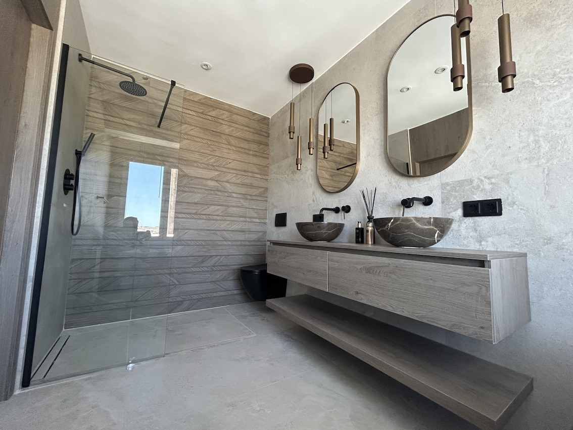 Modernes Badezimmer mit Doppelwaschtisch aus Stein, schwarzen Armaturen, ovalen Spiegeln und begehbarer Dusche mit Fliesen in Holzoptik.