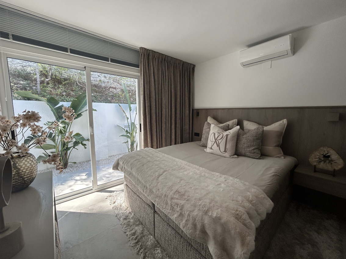 Modernes Schlafzimmer mit Holz-Kopfteil, Klimaanlage und Glasschiebetüren, die zu einer privaten Terrasse mit tropischen Pflanzen führen.