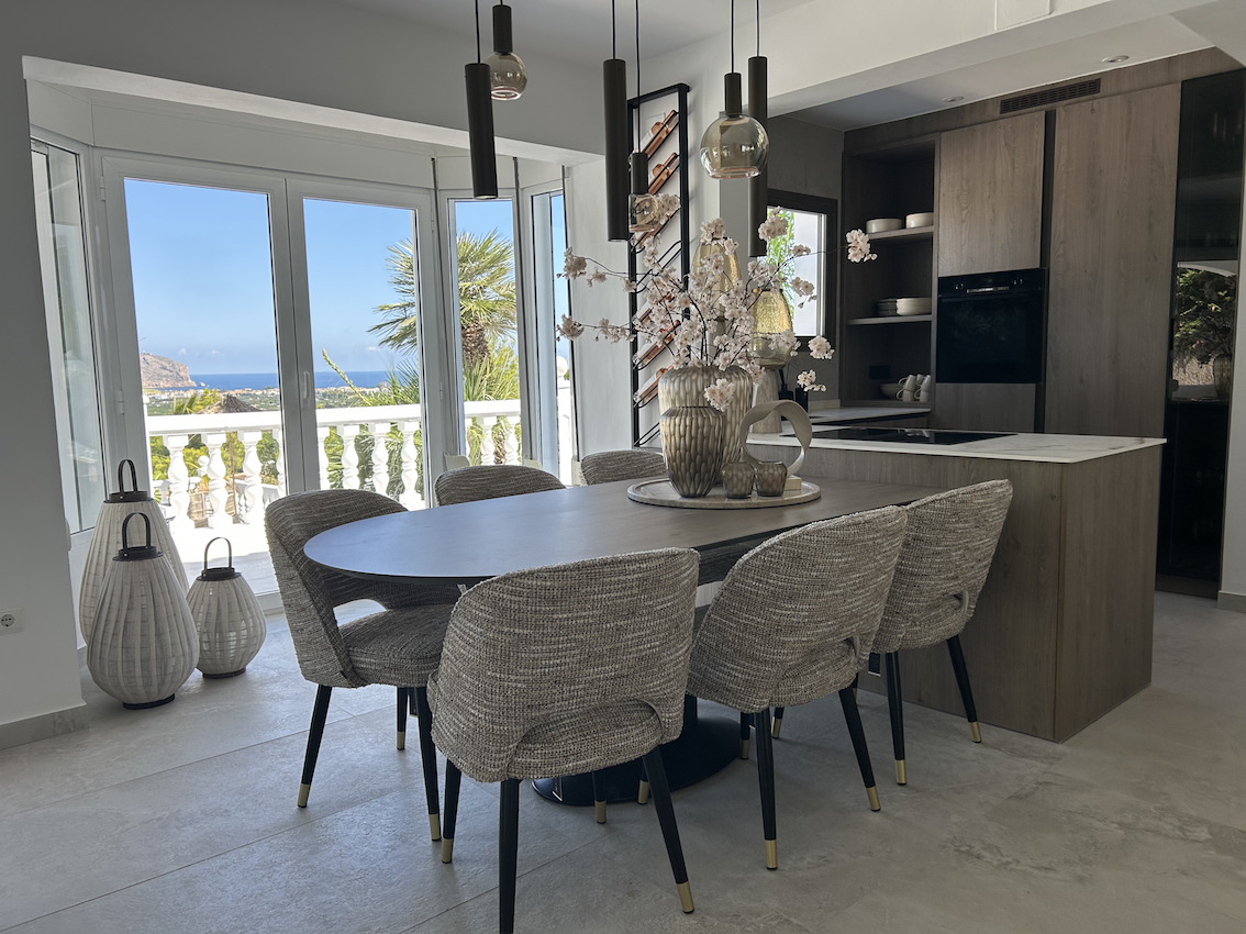 Offener Essbereich mit ovalem Tisch und integrierter Designerküche aus Holz, mit Panoramablick auf das Meer durch große Fensterfronten.