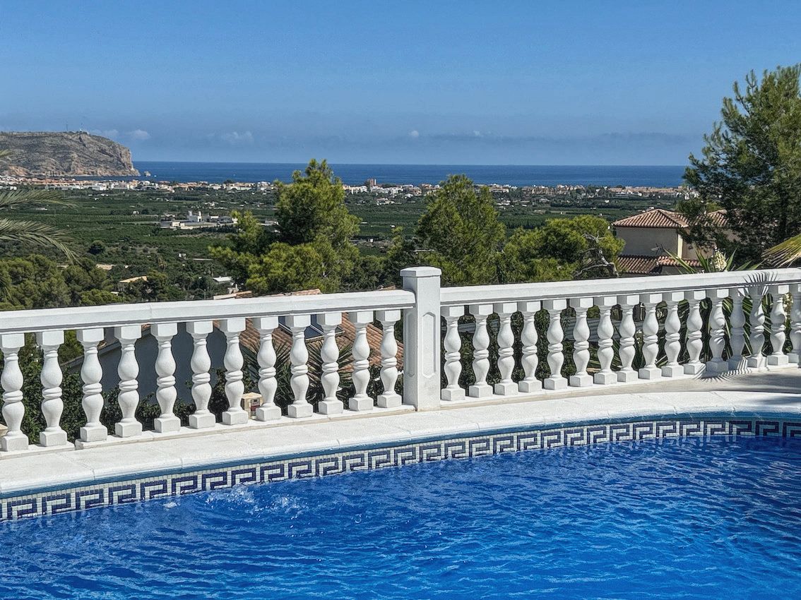 Schwimmbecken mit blauen Mosaikfliesen und weißer Balustrade, das einen Panoramablick auf das Mittelmeer, das Tal und die Küstenlinie bietet.