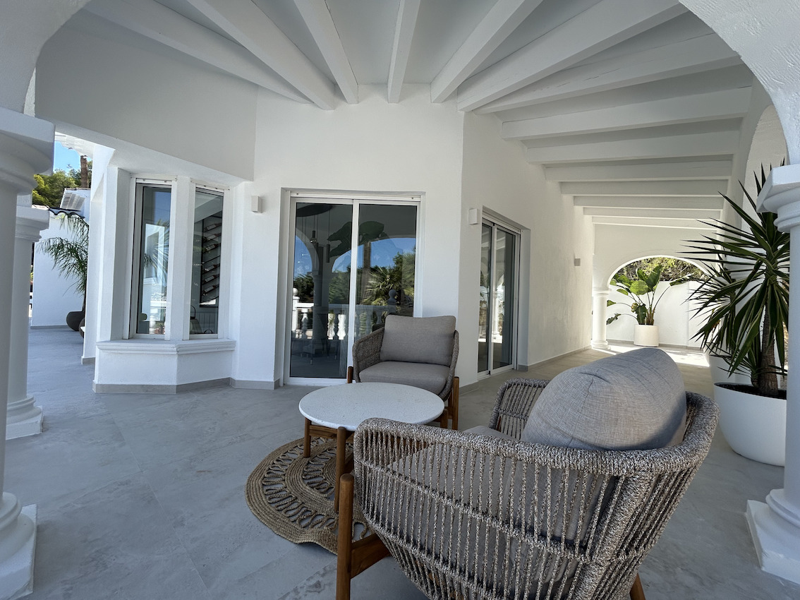 Überdachte Terrasse mit weiß gestrichenen Deckenbalken, modernen Korbsesseln und großformatigen Bodenfliesen, die direkt ins Haus führen.