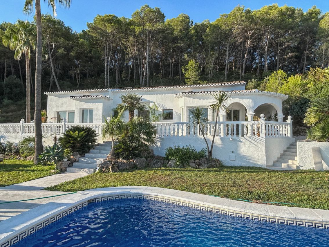 Weiße Villenfassade mit Balustradenterrasse, mediterranem Garten und Pool im Vordergrund, eingebettet in einen dichten Pinienwald.