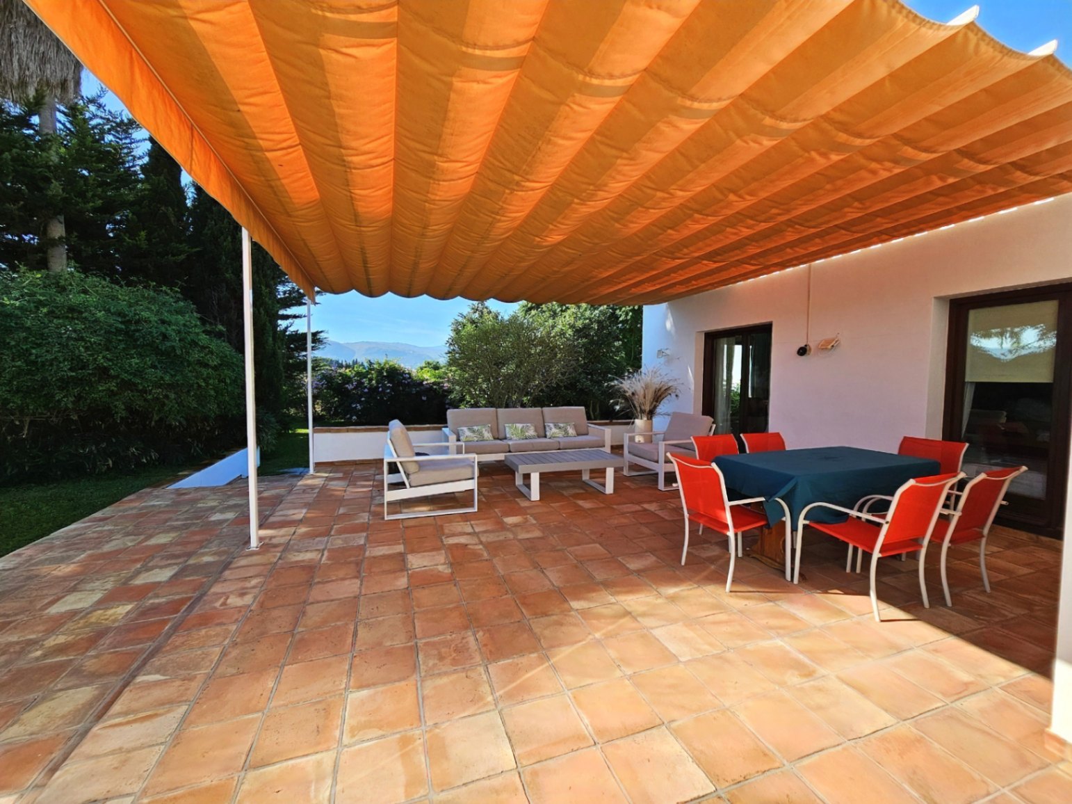 Außen-Terrasse mit handgefertigten Tonfliesen und einer großen orangefarbenen Markise. Mit Essbereich, Lounge und Blick auf den gepflegten Garten.