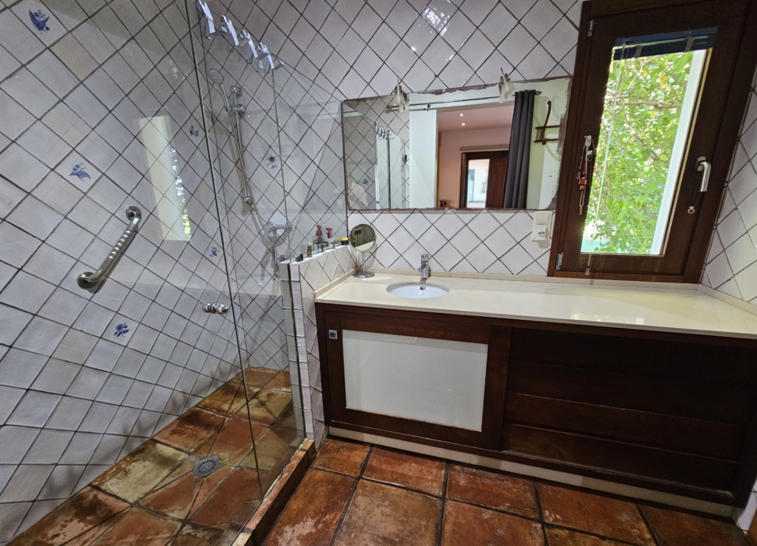 Badezimmer mit Glasdusche, handbemalten Wandfliesen und rustikalen Tonböden. Ein dunkler Holzwaschtisch und ein Fenster sorgen für natürliches Licht.