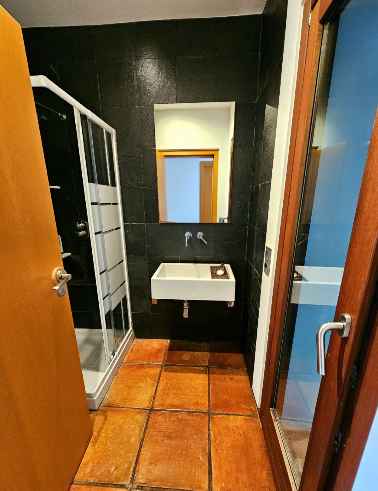 Badezimmer mit handgefertigten Terrakottafliesen, dunklen strukturierten Wandfliesen, modernem Hängewaschbecken und einer Duschkabine.