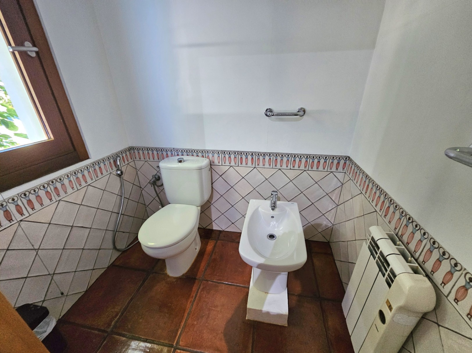 Badezimmerbereich mit WC und Bidet, ausgestattet mit handgefertigten Tonfliesen und dekorativen, handbemalten Bordüren an den Wänden.