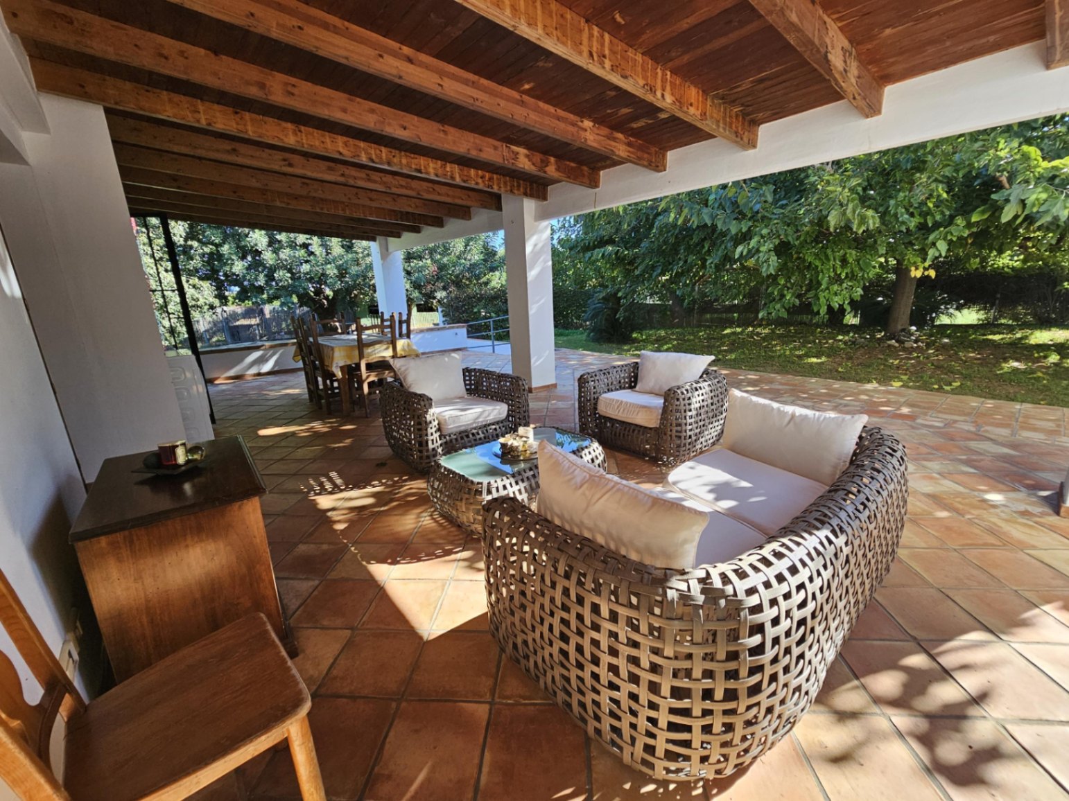 Große überdachte Terrasse mit Sichtbalkendecke, handgefertigten Tonfliesen und Korbgeflecht-Möbeln mit Blick in den weitläufigen Garten.