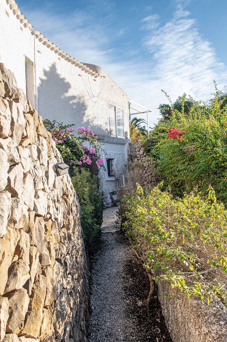 Außenpfad mit traditionellen Natursteinmauern und mediterraner Bepflanzung, der entlang der weiß getünchten Fassade der Villa führt.