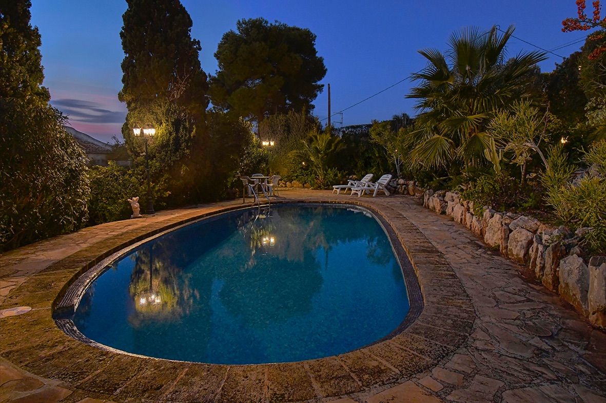 Beleuchteter Außenpool in der Abenddämmerung, umgeben von Natursteinpflaster und einem mediterranen Garten mit Palmen und Zypressen.