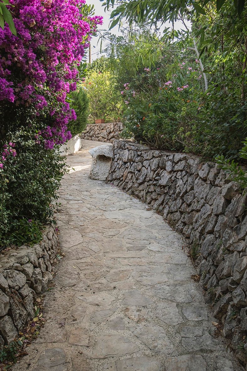 Ein Gartenweg aus Naturstein, gesäumt von traditionellen Trockenmauern und üppiger mediterraner Bepflanzung mit blühender Bougainvillea.