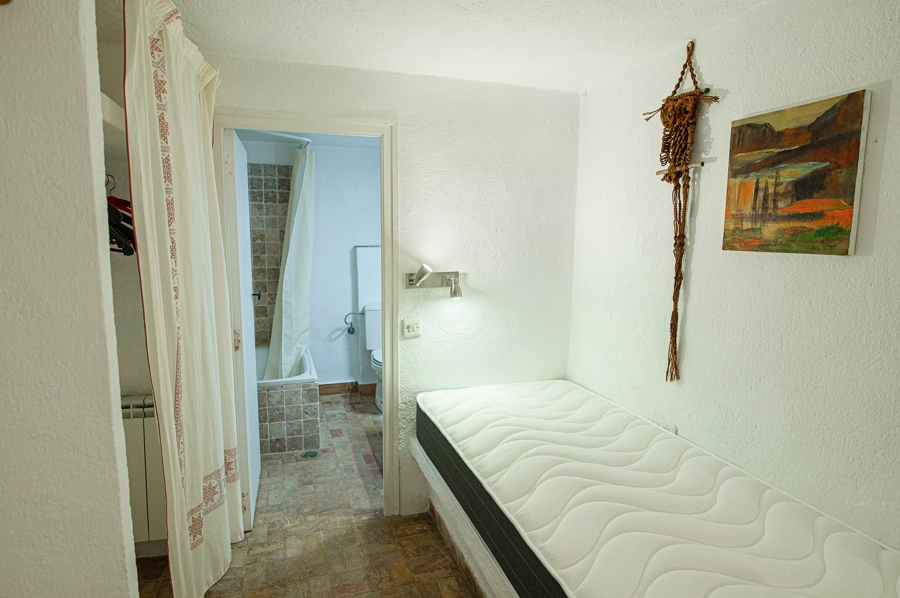 Funktionales Schlafzimmer mit Einzelbett auf gemauertem Sockel, weißen Texturwänden und En-Suite-Bad mit rustikalen Fliesen in Erdtönen.