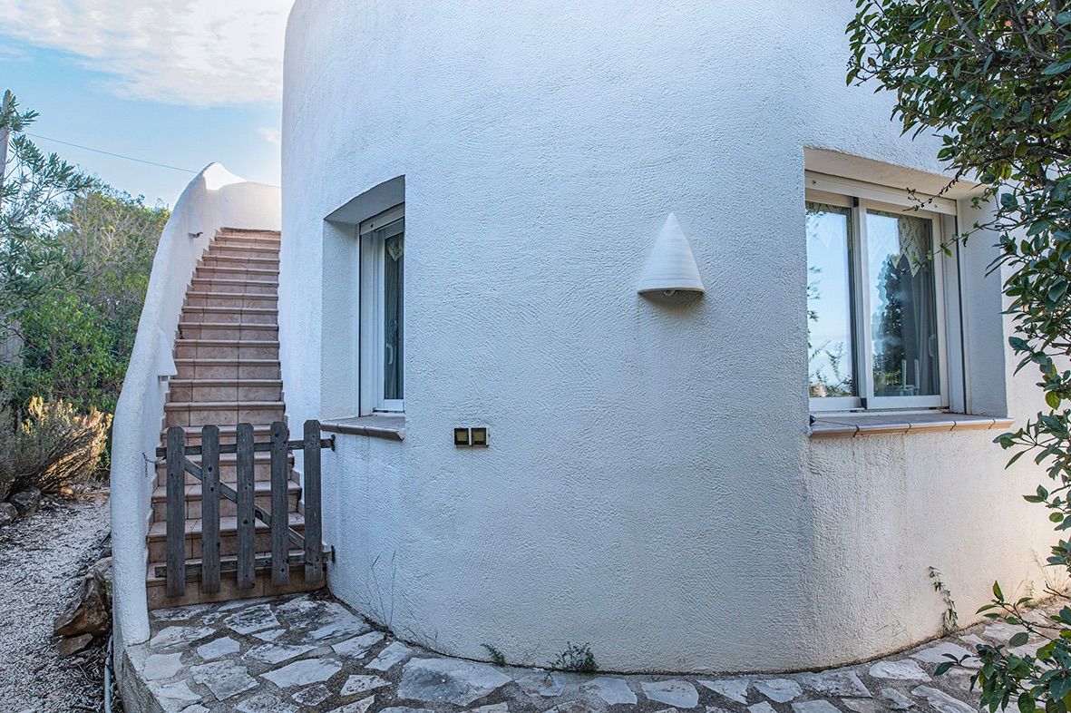 Geschwungene mediterrane Fassade mit weißem Außenputz, einer Außentreppe mit Keramikstufen und einem kleinen Holztor am Zugang.