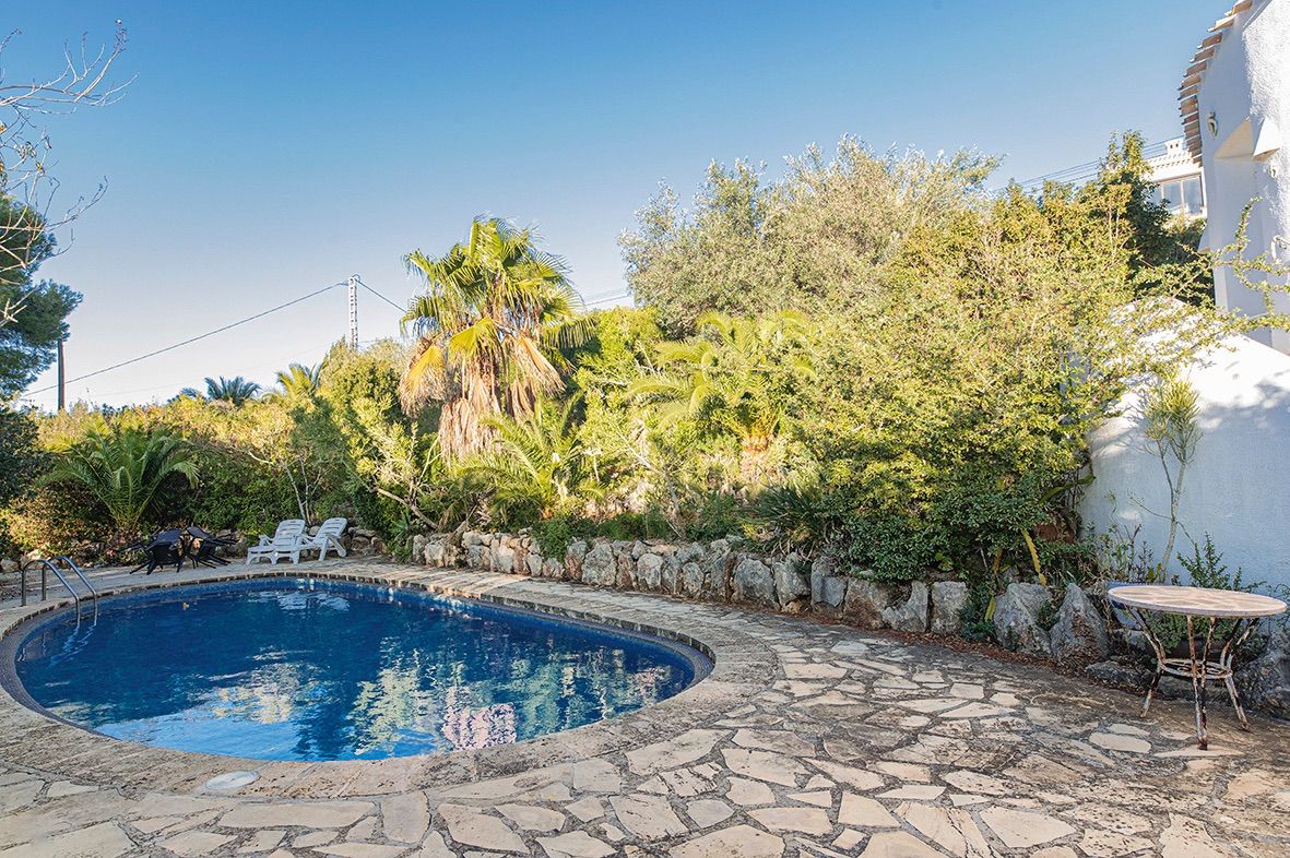 Nierenförmiger Pool mit Natursteinterrasse, rustikalen Steinmauern und einem üppigen mediterranen Garten mit Palmen unter blauem Himmel.