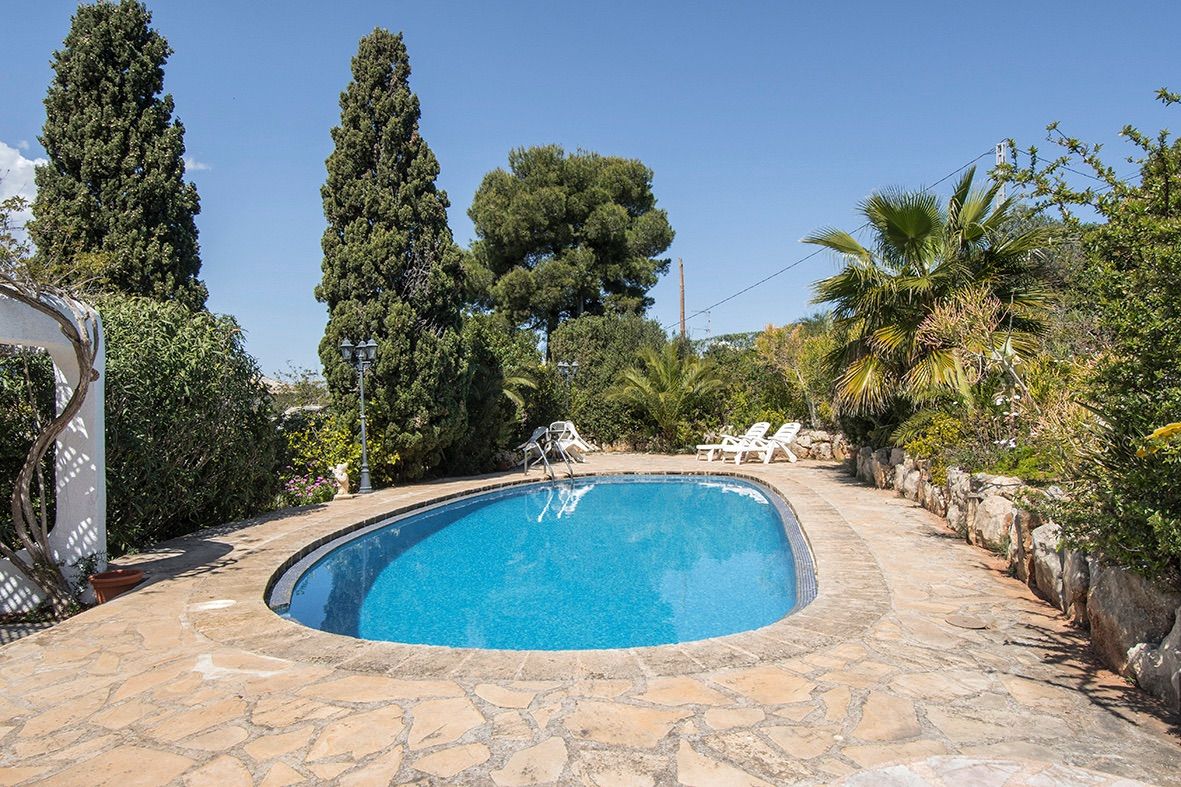 Ovaler Swimmingpool mit Natursteinterrasse, umgeben von einem mediterranen Garten mit hohen Zypressen und Palmen unter blauem Himmel.