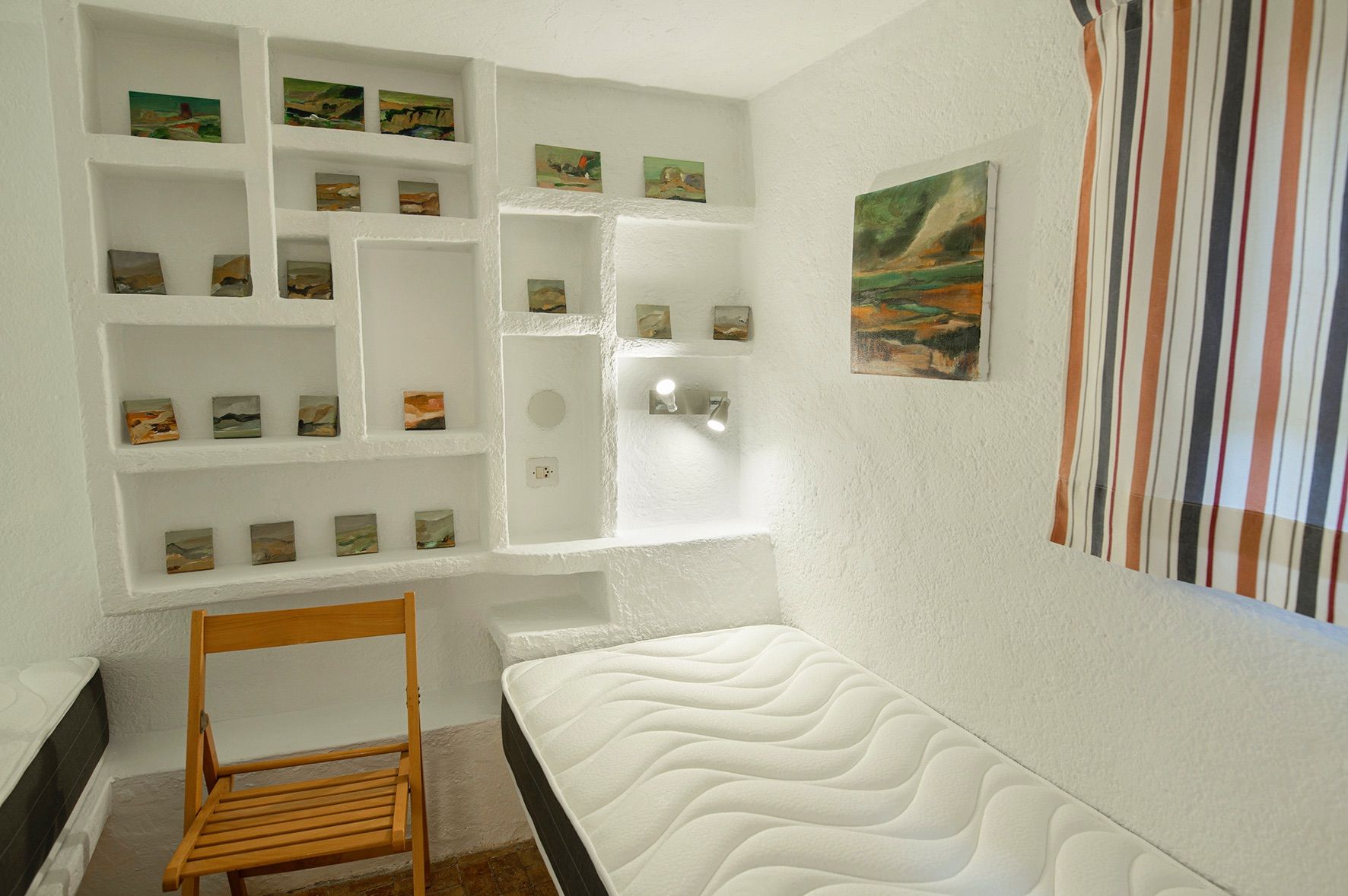 Schlafzimmer mit weiß verputzten Wänden und integrierten Wandnischen für Kunstwerke, ausgestattet mit einer Einzelmatratze und einem Holzstuhl.