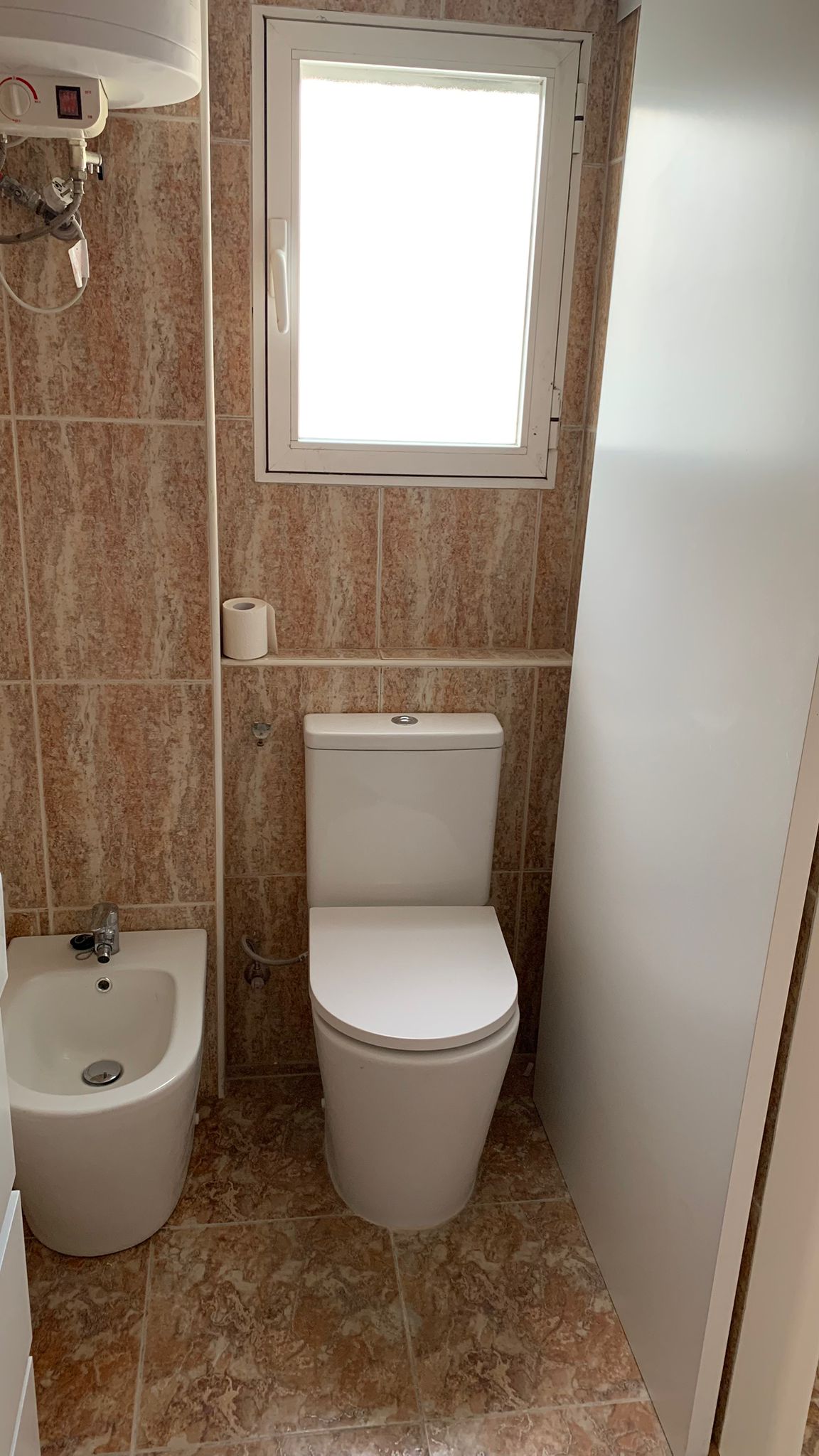 Badezimmer mit weißem WC und Bidet, erdfarbenen Fliesen in Marmoroptik und einem Fenster für natürliches Licht und Belüftung.