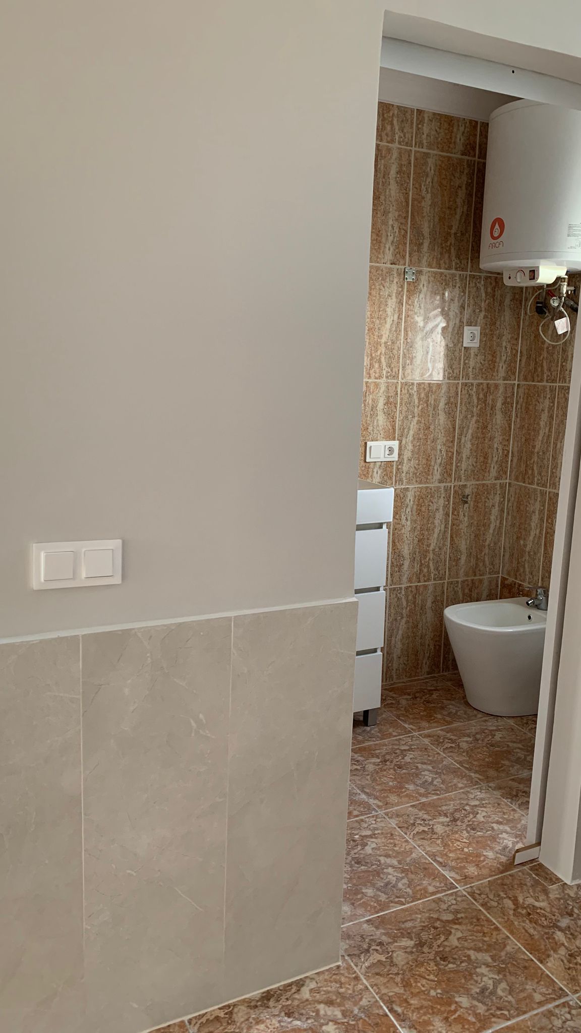 Blick in das Badezimmer mit Bidet, weißem Unterschrank und Elektro-Warmwasserspeicher, ausgestattet mit braunen Fliesen in Marmoroptik.