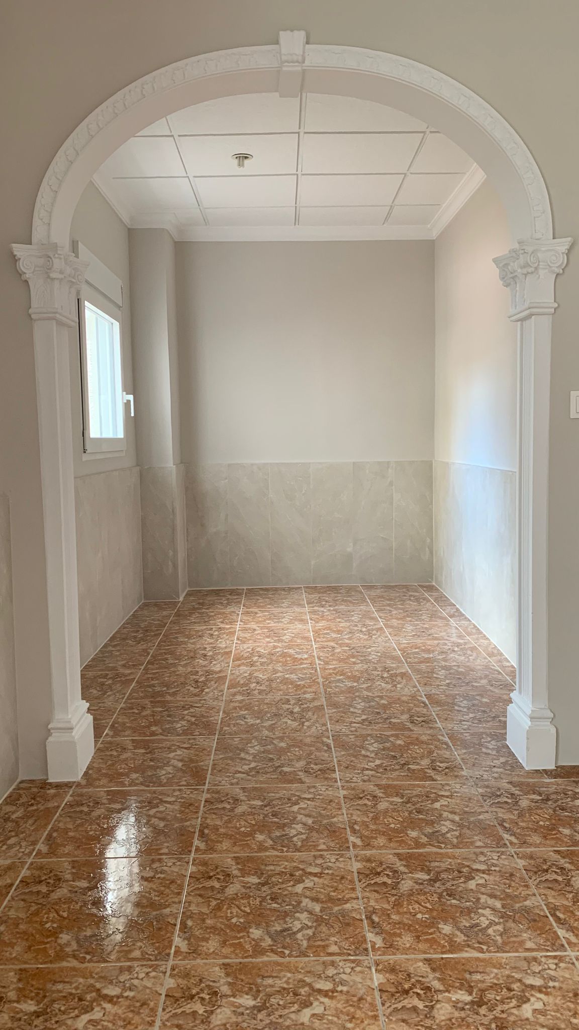 Eingangsbereich mit klassischem Dekorbogen, erdfarbenen Keramikfliesen und grauem Fliesensockel. Ein Seitenfenster sorgt für natürliches Licht im Raum.