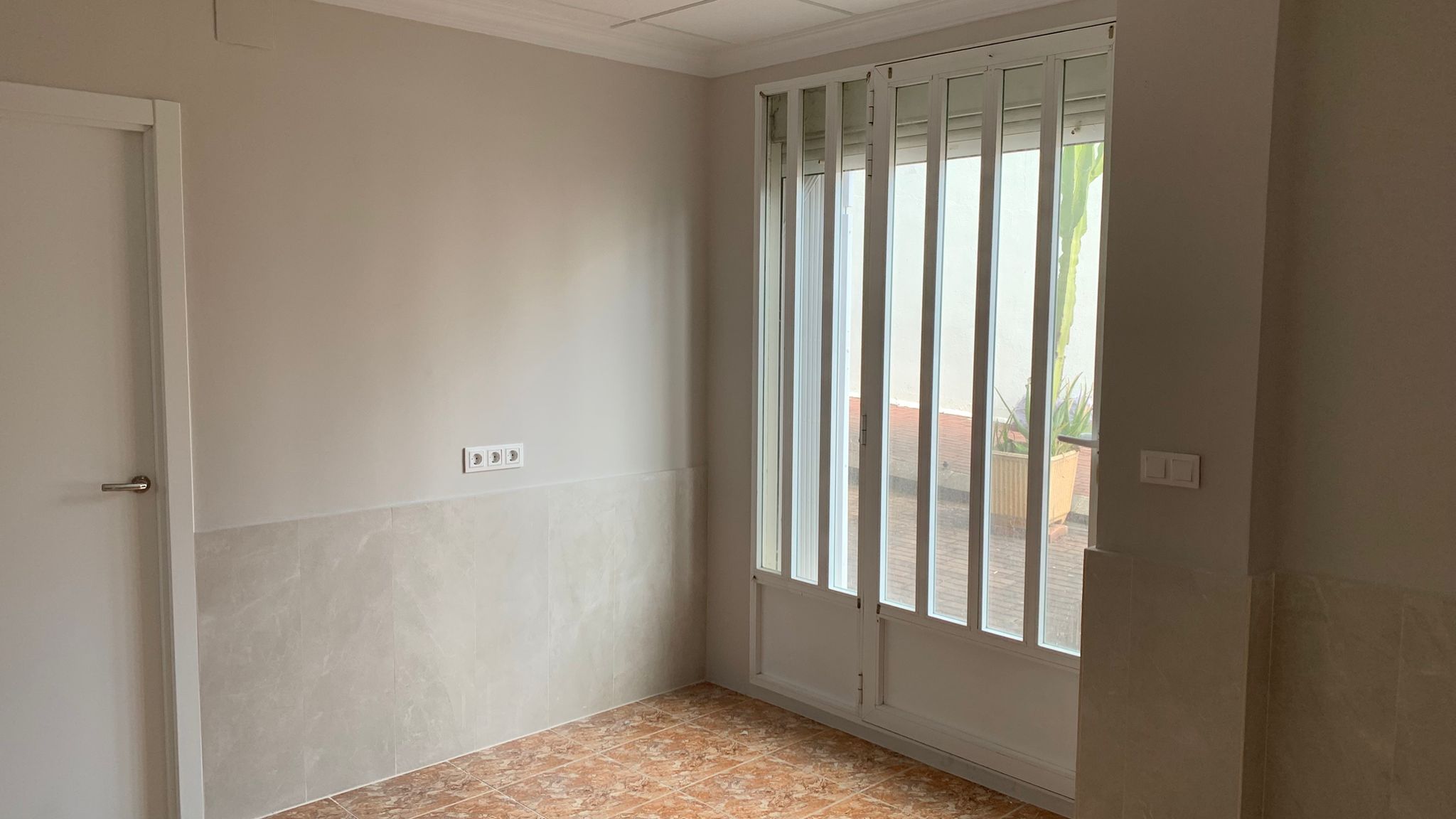 Innenraum mit Terrakotta-Bodenfliesen, Wandverkleidung in Marmoroptik und einer Glastür mit Sicherheitsgittern, die zum Außenbereich führt.