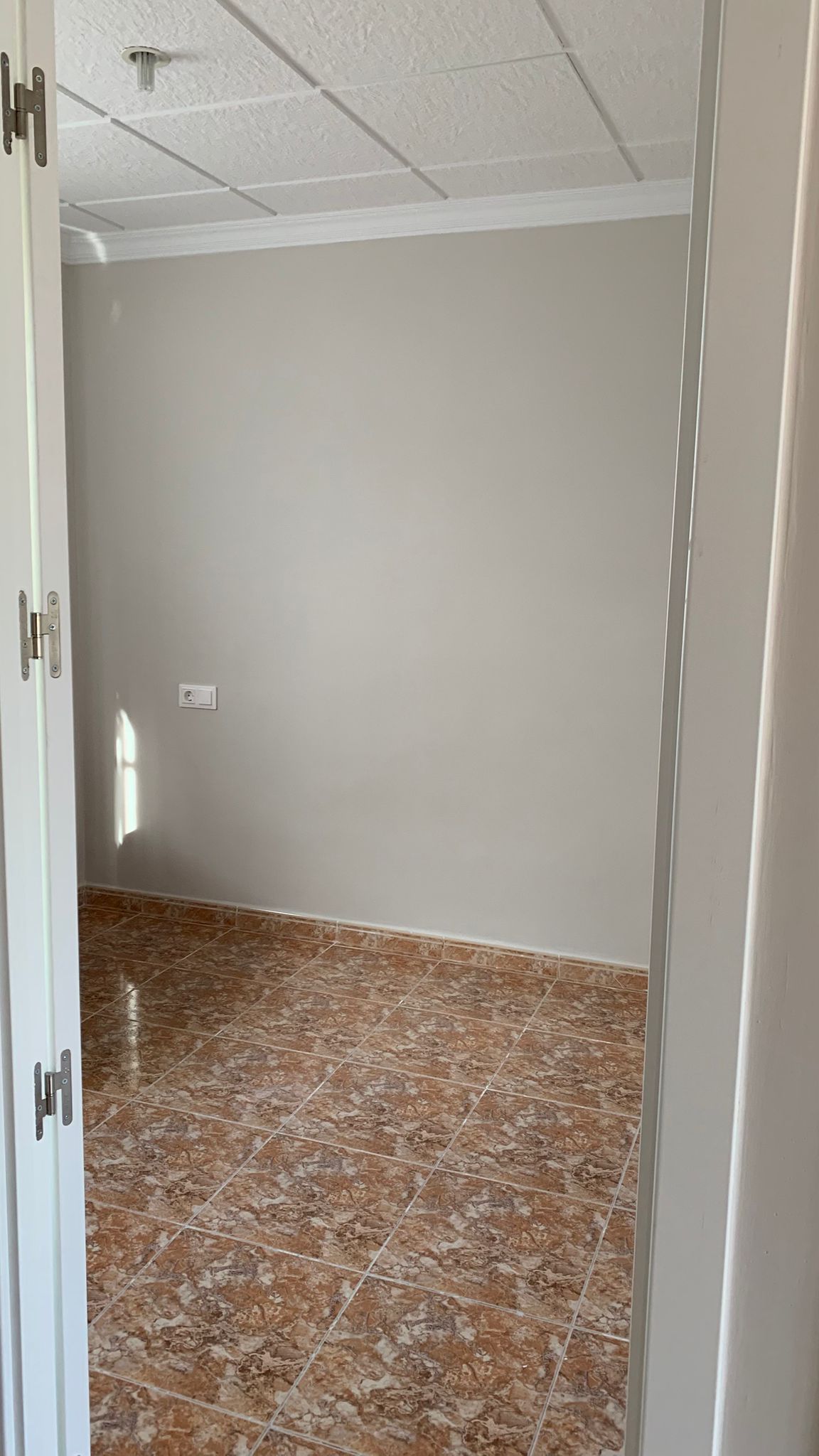 Leerer Raum mit braunen Keramikfliesen, hellgrauen Wänden und einer weißen Rasterdecke. Seitlicher Lichteinfall betont die schlichte und gepflegte Gestaltung.