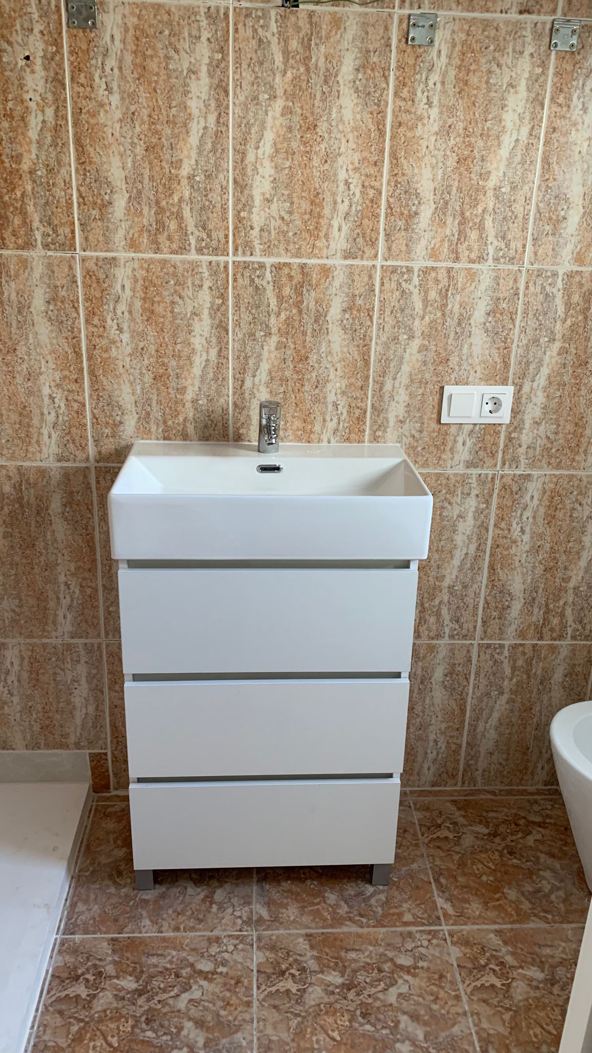Moderner weißer Badezimmer-Waschtisch mit drei Schubladen und integriertem Waschbecken, umgeben von erdfarbenen Keramikfliesen.