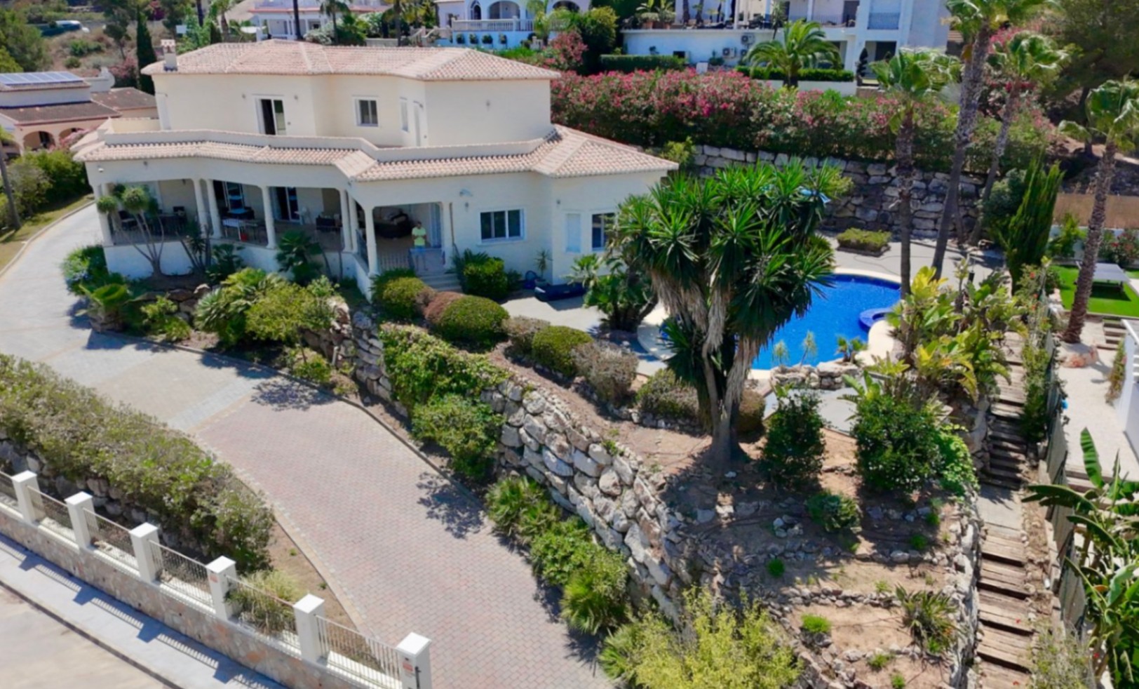 Außenansicht der Villa mit weißer Fassade, Ziegeldächern, großzügiger gepflasterter Einfahrt, Natursteinmauern und mediterran angelegten Gärten mit Pool.