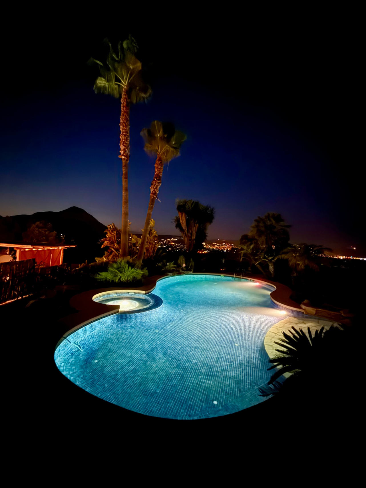 Beleuchteter Freiform-Pool mit integriertem Whirlpool bei Nacht, umgeben von Palmen mit Blick auf die Stadtlichter von Jávea und das Montgó-Massiv.