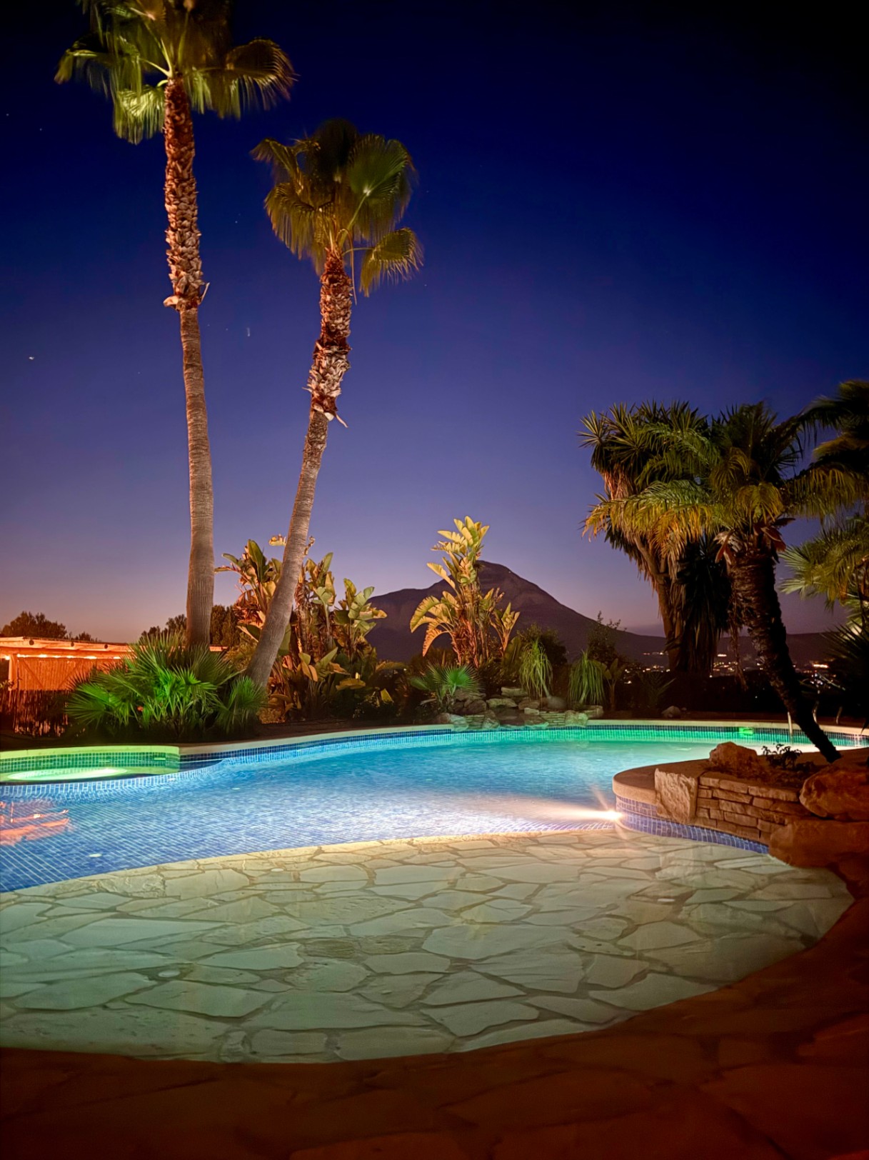 Beleuchteter Pool bei Nacht mit Natursteinpflasterung und Blick auf die Silhouette des Montgó, umgeben von Palmen und tropischer Bepflanzung.