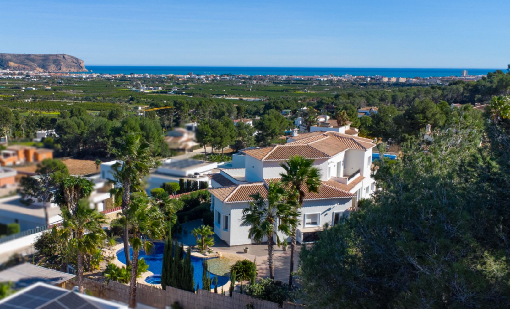 Erhöhte Ansicht der weißen Villa mit Ziegeldächern, organisch geformtem Pool und mediterranem Garten mit weitem Blick auf die Küste von Jávea.