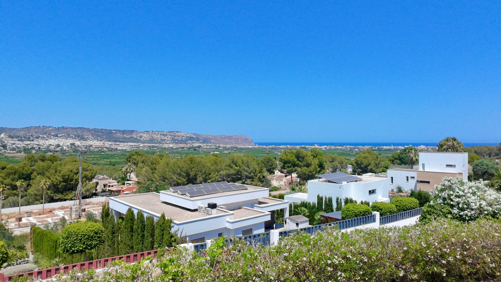 Erhöhter Panoramablick auf das Mittelmeer und das Tal, mit modernen weißen Villen, Solaranlagen auf den Dächern und üppiger mediterraner Vegetation.