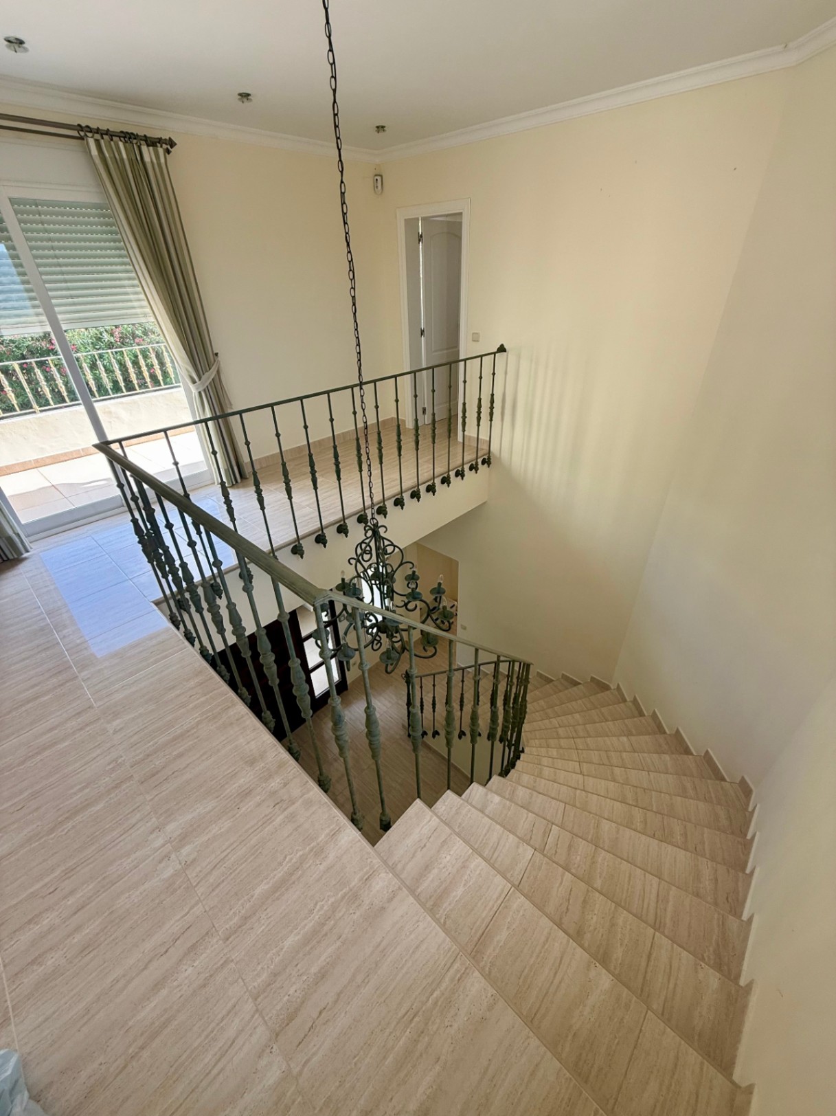 Geschwungene Marmortreppe mit schmiedeeisernem Geländer und zentralem Kronleuchter, die zu einem hellen Flur mit Terrassenzugang führt.