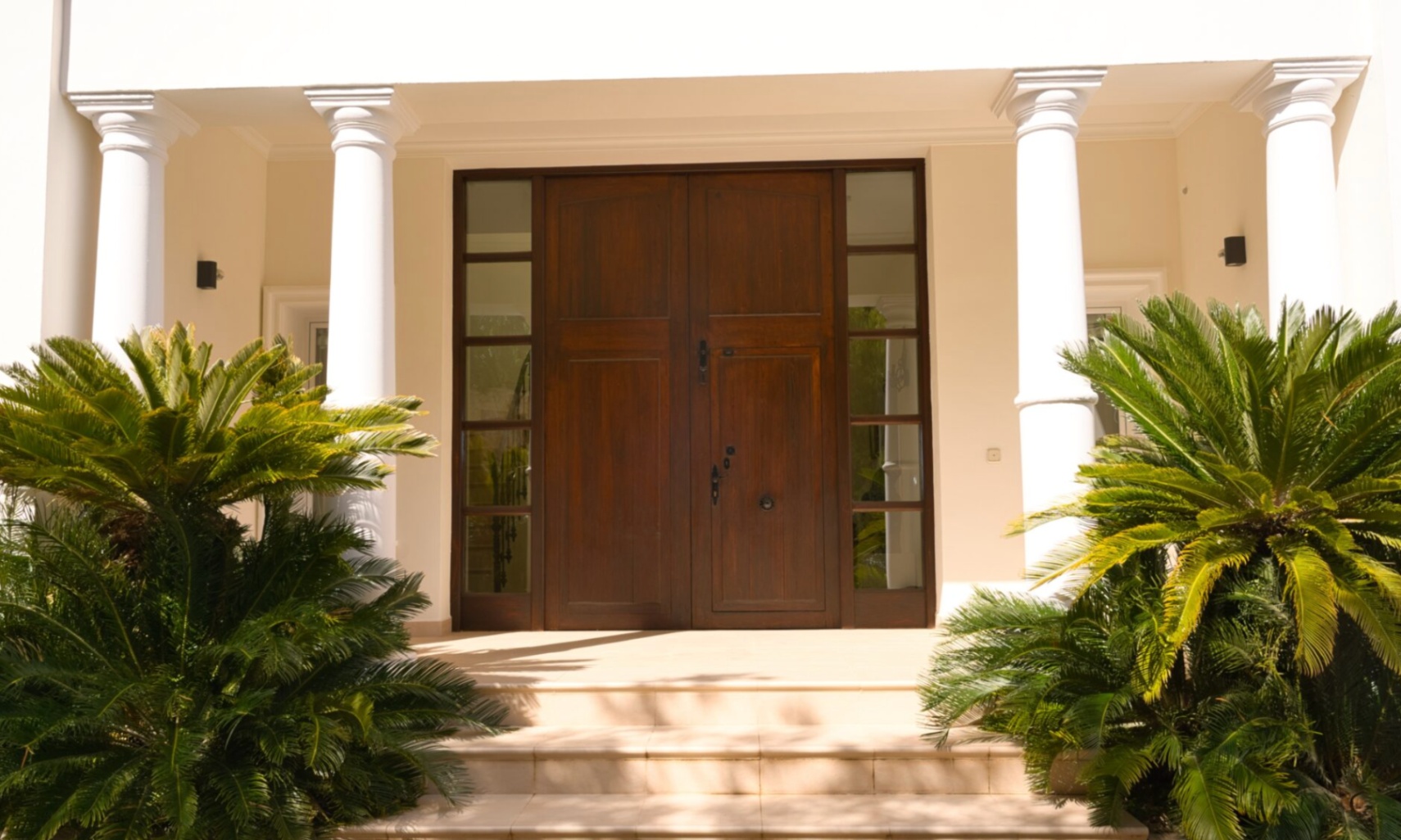 Haupteingang mit massiver Doppeltür aus dunklem Holz, flankiert von klassischen weißen Säulen und üppiger mediterraner Bepflanzung.