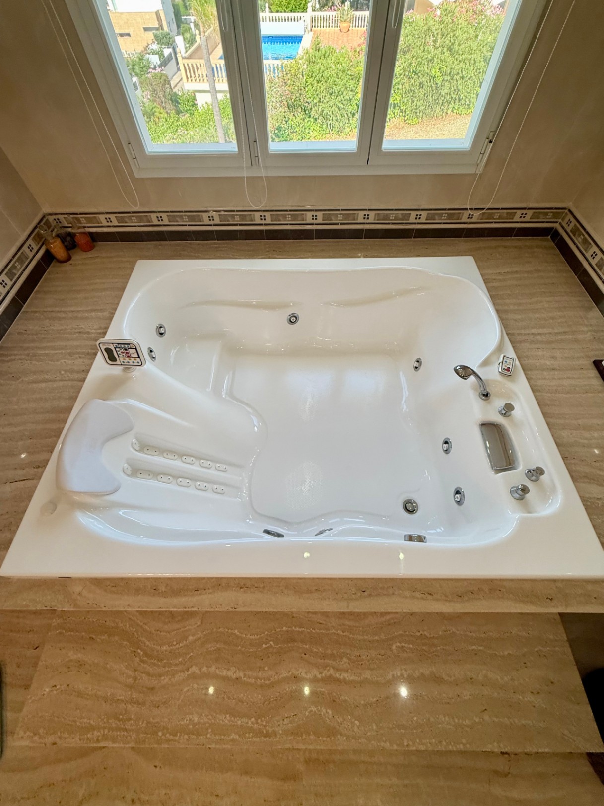 In Travertin-Marmor eingefasste Hydromassage-Badewanne mit digitaler Steuerung und Kopfstütze, platziert vor einem Fenster mit Blick auf den Garten und Poolbereich.