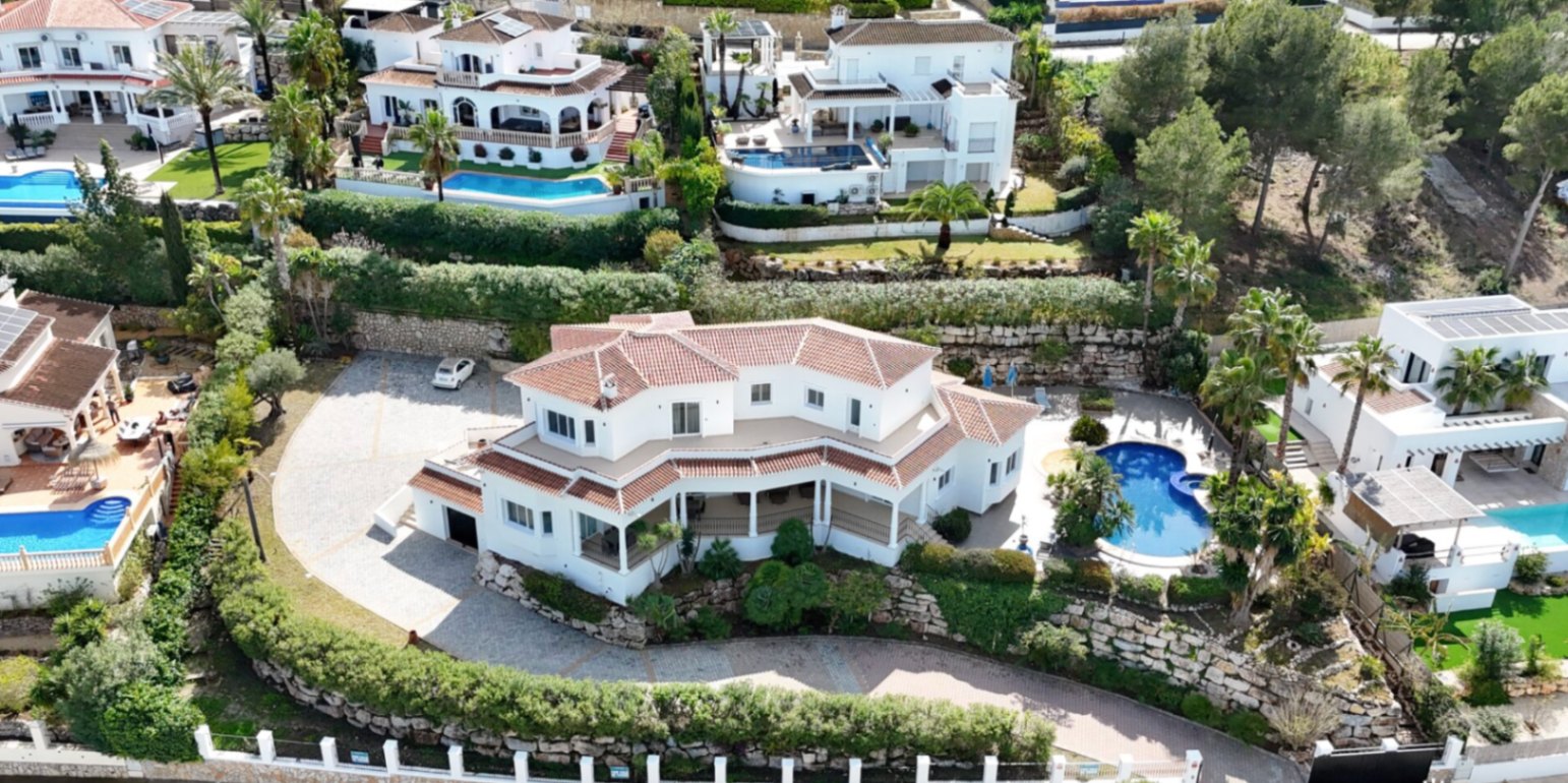 Luftaufnahme der Villa mit weißer Fassade und Ziegeldach, großzügigem gepflastertem Parkplatz, Natursteinmauern und privatem Pool inmitten mediterraner Gärten.
