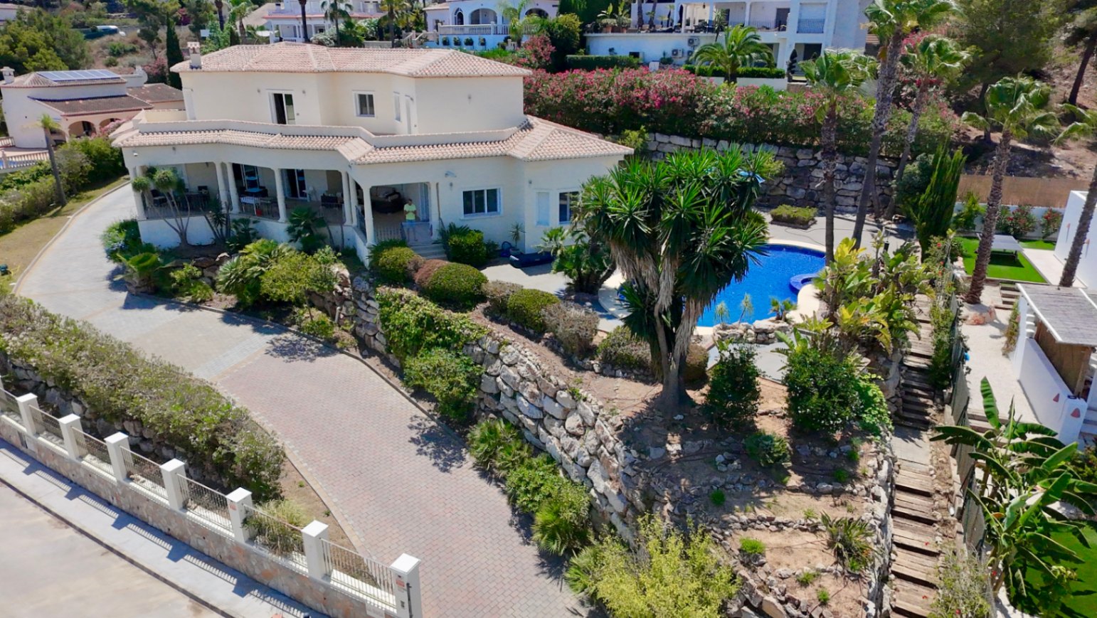 Luftbild der Villa mit zweistöckiger Architektur, großzügiger gepflasterter Auffahrt und einem Poolbereich, der in terrassierte Gärten mit Natursteinmauern eingebettet ist.