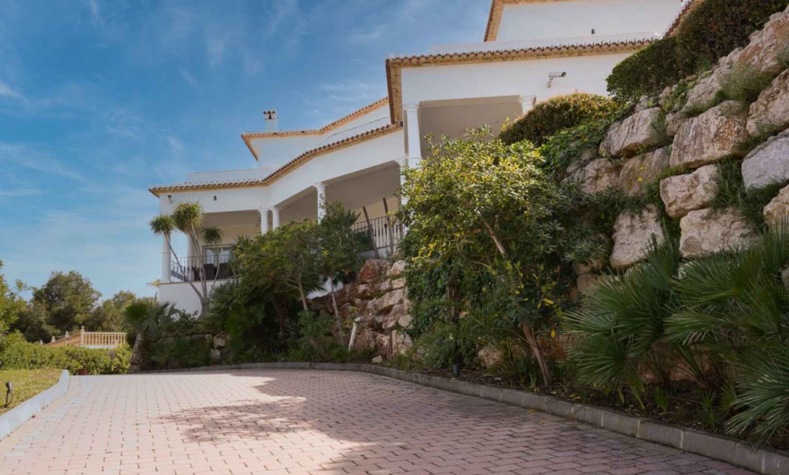 Mediterrane Außenansicht mit weißen Säulen, überdachten Terrassen und einer gepflasterten Auffahrt, gesäumt von einer Natursteinmauer und lokaler Bepflanzung.