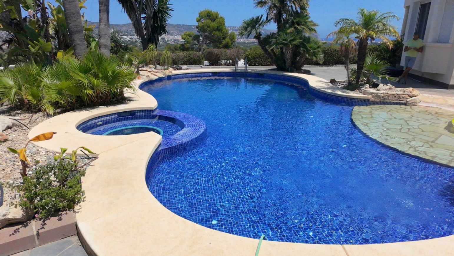 Organisch geformter Außenpool mit blauen Mosaikfliesen, integriertem Whirlpool und Natursteinbereich, umgeben von Palmen und mediterraner Bepflanzung.