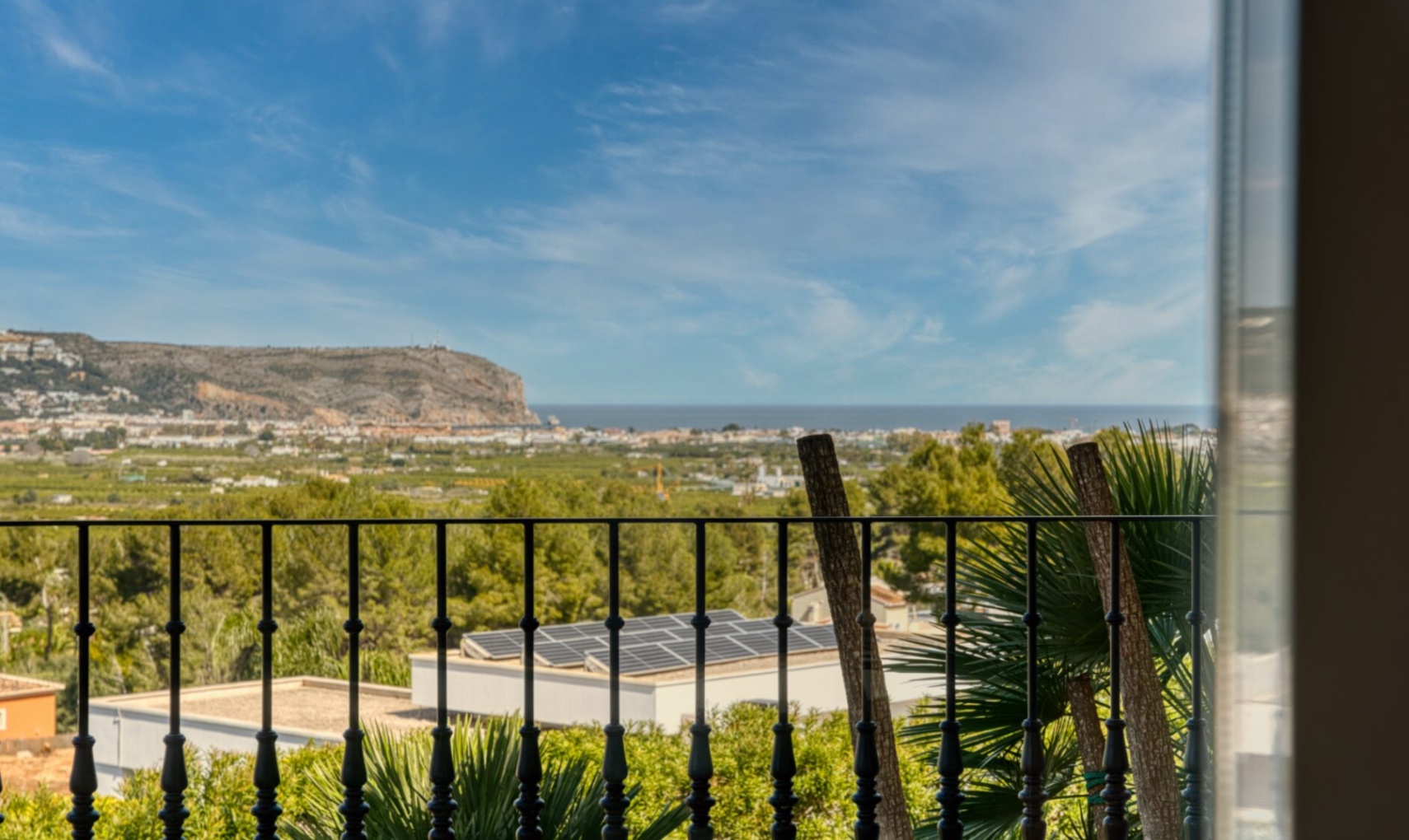 Panoramablick von der Terrasse mit Schmiedeeisengeländer auf das Mittelmeer und das Kap San Antonio unter strahlend blauem Himmel.