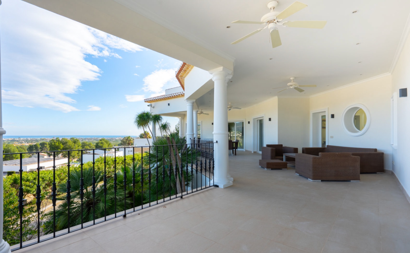 Überdachte Terrasse mit weißen Säulen, Deckenventilatoren und Rattan-Lounge-Möbeln mit Panoramablick auf das Mittelmeer und die umliegende Landschaft.