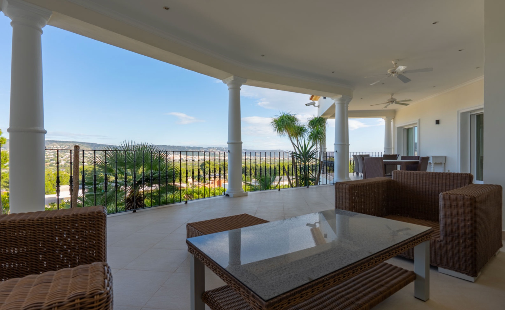 Überdachte Terrasse mit weißen Säulen, Korbmöbeln und Panoramablick auf das Tal. Die Decke ist mit Ventilatoren und Einbaustrahlern ausgestattet.