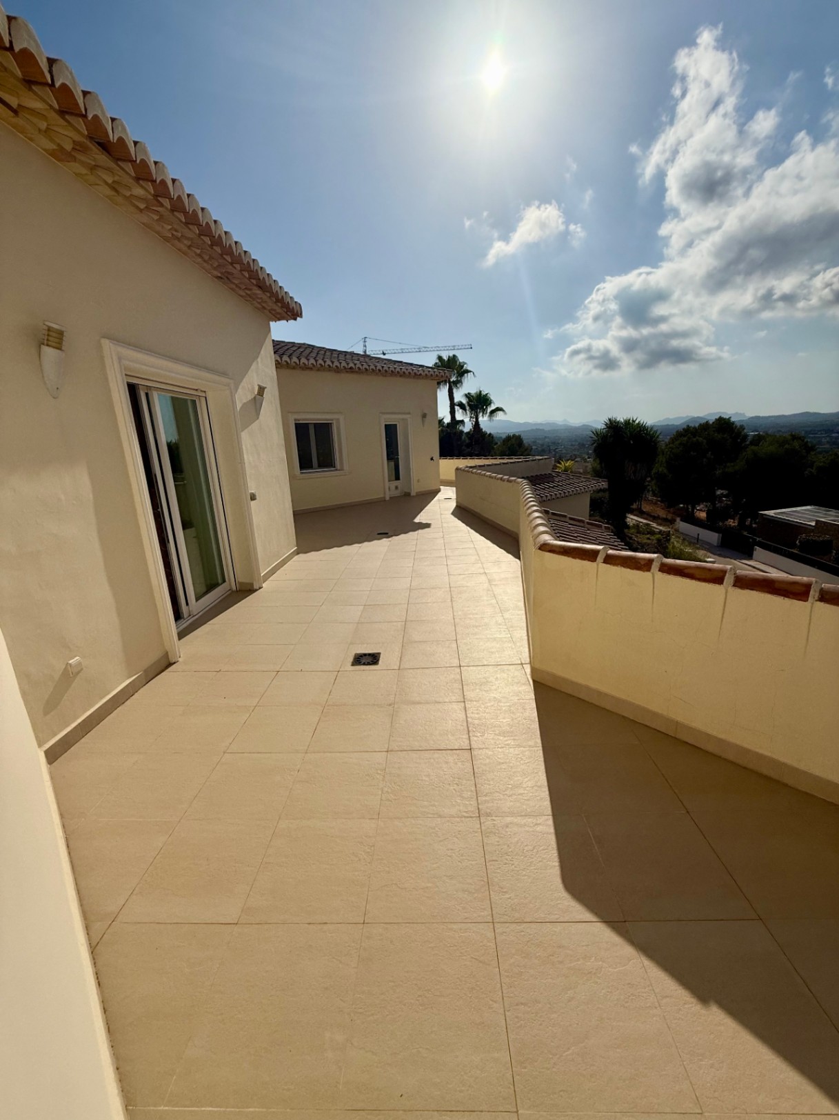 Weitläufige Oberterrasse mit beigen Bodenfliesen und Panoramablick auf das Tal und die Berge, zugänglich über große Glasschiebetüren bei hellem Tageslicht.